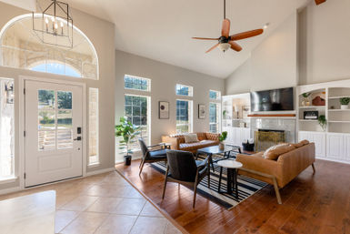 Sunlit modern living room and foyer with arched transom window, vaulted ceiling and fan, leather sofas, black accent chairs, striped rug, fireplace with TV above, built-in white shelving and hardwood floors.