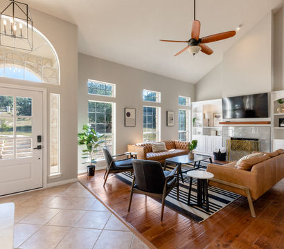 Sunlit modern living room and foyer with arched transom window, vaulted ceiling and fan, leather sofas, black accent chairs, striped rug, fireplace with TV above, built-in white shelving and hardwood floors.