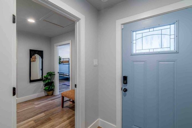 Cheerful home entryway with a light-blue front door featuring decorative frosted glass and a keypad lock; hardwood floors lead to a hallway with a black-framed mirror, potted plant, woven bench, and a glimpse of a bunk bed in the adjacent room.