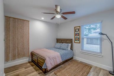 Cozy modern farmhouse-style bedroom with wooden platform bed dressed in blue floral sheets and pink quilt, ceiling fan, hardwood floors, woven area rug, floor lamp, window with blinds, and neutral walls with framed floral art.
