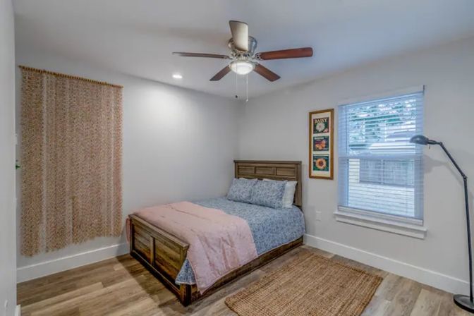 Cozy modern farmhouse-style bedroom with wooden platform bed dressed in blue floral sheets and pink quilt, ceiling fan, hardwood floors, woven area rug, floor lamp, window with blinds, and neutral walls with framed floral art.
