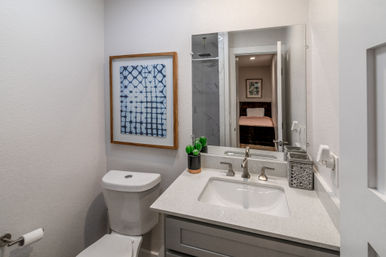 Modern small residential bathroom with white quartz vanity, undermount sink, brushed-nickel faucet, toilet, framed blue wall art and small succulent, mirror reflecting bedroom and shower.