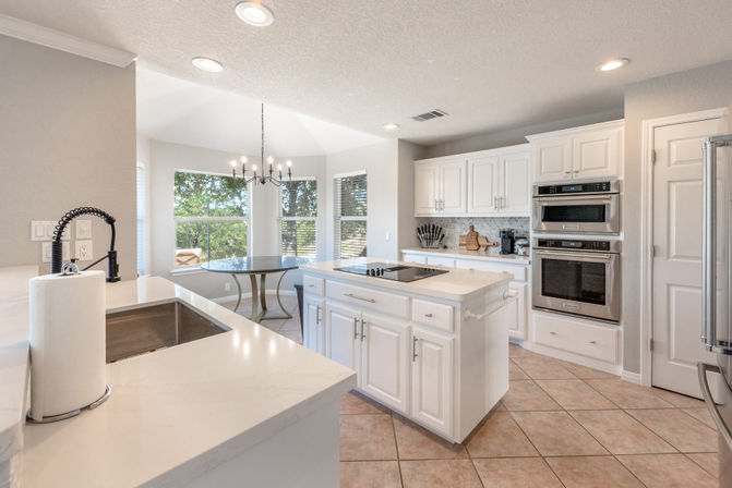 Bright modern white kitchen with central island and cooktop, quartz countertops, stainless steel double wall ovens, farmhouse sink, tile floor and sunny bay-window breakfast nook with chandelier