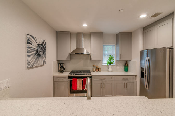 Sleek modern kitchen with light gray cabinetry, quartz island and faucet in foreground, stainless steel fridge and gas range with vent hood, red dish towels and a small window plant