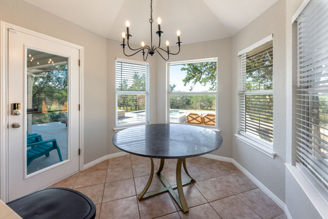 Sunny breakfast nook with round marble table and black iron chandelier, bay windows and glass door overlooking patio, pool and trees