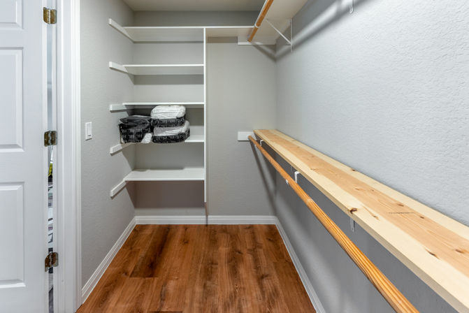 Bright walk-in closet with wood-look flooring, white built-in shelves holding folded linens, long wooden hanging rods and neutral gray walls.