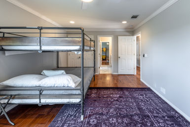 Modern residential bedroom interior with black metal bunk bed and white bedding, hardwood floors, patterned purple area rug, light gray walls, recessed ceiling lights and open doorway to hallway and bathroom