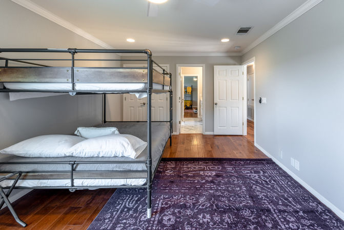 Modern residential bedroom interior with black metal bunk bed and white bedding, hardwood floors, patterned purple area rug, light gray walls, recessed ceiling lights and open doorway to hallway and bathroom