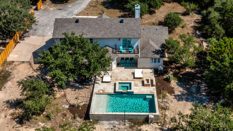 Aerial drone view of a modern two-story home with a stone backyard patio, sparkling rectangular pool and attached spa, turquoise patio chairs on a balcony, and surrounding oak trees.