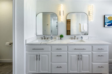 Bright modern white double-sink bathroom vanity with marble countertop, two arched mirrors, chrome faucets, vertical glass wall sconces and a small potted plant.