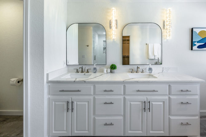 Bright modern white double-sink bathroom vanity with marble countertop, two arched mirrors, chrome faucets, vertical glass wall sconces and a small potted plant.