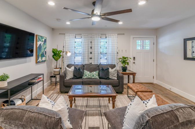 Cozy modern living room with a gray sofa, wooden coffee table, wall-mounted TV, ceiling fan, indoor plants, striped rug and a white front door.