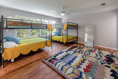 Sunlit modern bedroom with two metal twin-over-full bunk beds dressed in yellow scalloped quilts, large windows overlooking trees, colorful geometric area rug on hardwood floors, ceiling fan and full-length mirror.