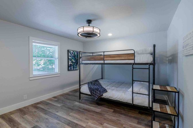 Cozy modern guest bedroom with metal-frame double-over-double bunk bed with wood accents, white bedding, wood-look plank floors, round ceiling light, window with blinds and abstract wall art.
