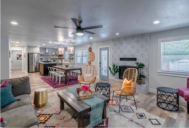 Open-concept living room and kitchen with patterned tile fireplace wall, rattan chairs, wooden coffee table, dining table on a purple rug, stainless steel refrigerator, pendant lights, ceiling fan and colorful boho accents.