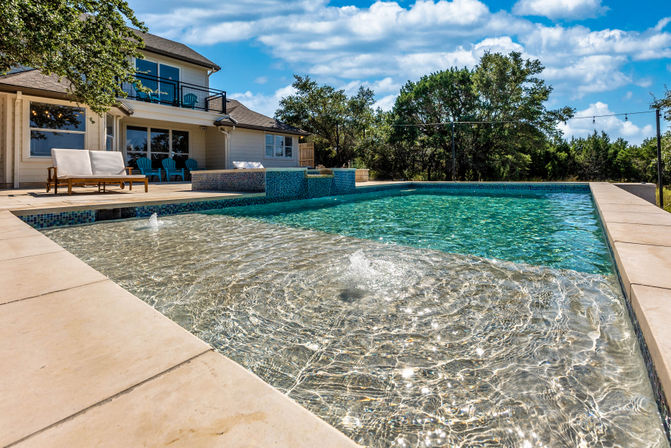Modern suburban backyard swimming pool with shallow sun shelf and bubbling jets, turquoise water, stone pool deck, lounge seating and a two-story house framed by trees under a blue sky