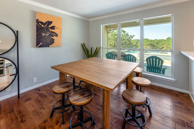 Cozy sunlit dining nook with a large wooden table and round industrial-style stools on hardwood floors, large window revealing patio chairs, a swimming pool, and treed yard outside.
