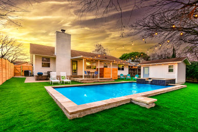 Backyard suburban home with rectangular pool, vivid green lawn, patio dining and lounge chairs, string lights at twilight and a small pool house.