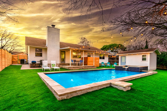 Backyard suburban home with rectangular pool, vivid green lawn, patio dining and lounge chairs, string lights at twilight and a small pool house.