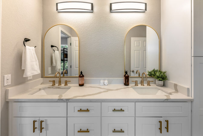 Bright modern double-sink bathroom vanity with white marble-look countertop, brass faucets and hardware, two arched mirrors, wall sconces, white cabinets, towel hook and small potted plant.