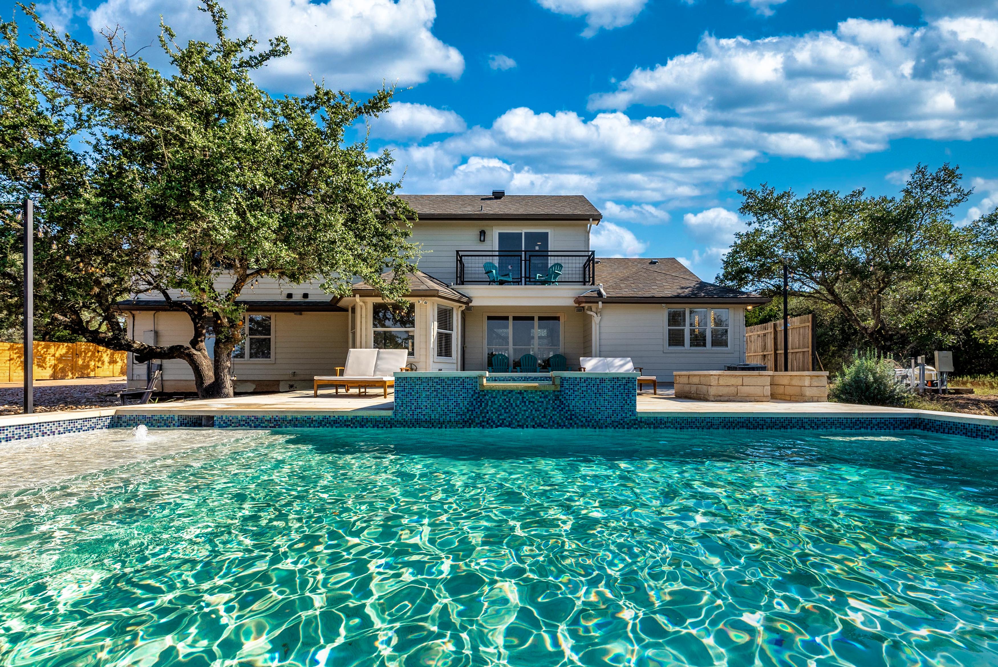 Sparkling backyard swimming pool in front of a two-story suburban home with patio, lounge chairs, oak trees and a bright blue sky