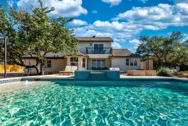 Sparkling backyard swimming pool in front of a two-story suburban home with patio, lounge chairs, oak trees and a bright blue sky