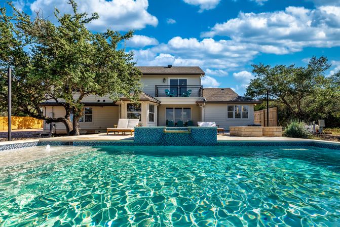 Sparkling backyard swimming pool in front of a two-story suburban home with patio, lounge chairs, oak trees and a bright blue sky