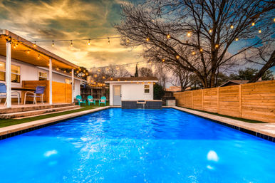 Vivid blue backyard pool at dusk with hanging string lights, covered patio and turquoise chairs beside a fenced residential yard and small pool house
