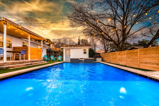 Vivid blue backyard pool at dusk with hanging string lights, covered patio and turquoise chairs beside a fenced residential yard and small pool house