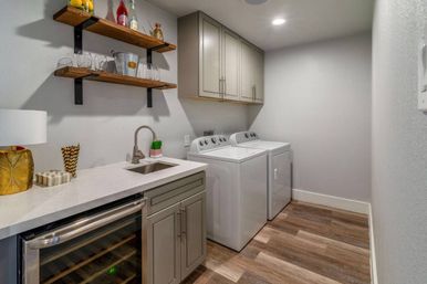 Bright modern laundry room with white top-load washer and dryer, gray shaker cabinets, quartz countertop with sink, built-in wine fridge, wood floating shelves with glassware, and wood-look flooring.