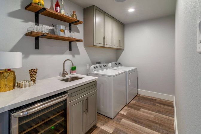Bright modern laundry room with white top-load washer and dryer, gray shaker cabinets, quartz countertop with sink, built-in wine fridge, wood floating shelves with glassware, and wood-look flooring.