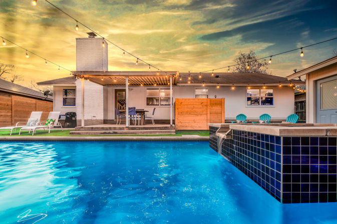 Sunset suburban backyard with an illuminated blue swimming pool and tiled spa, covered patio dining area, lounge chairs, turquoise Adirondacks and glowing string lights
