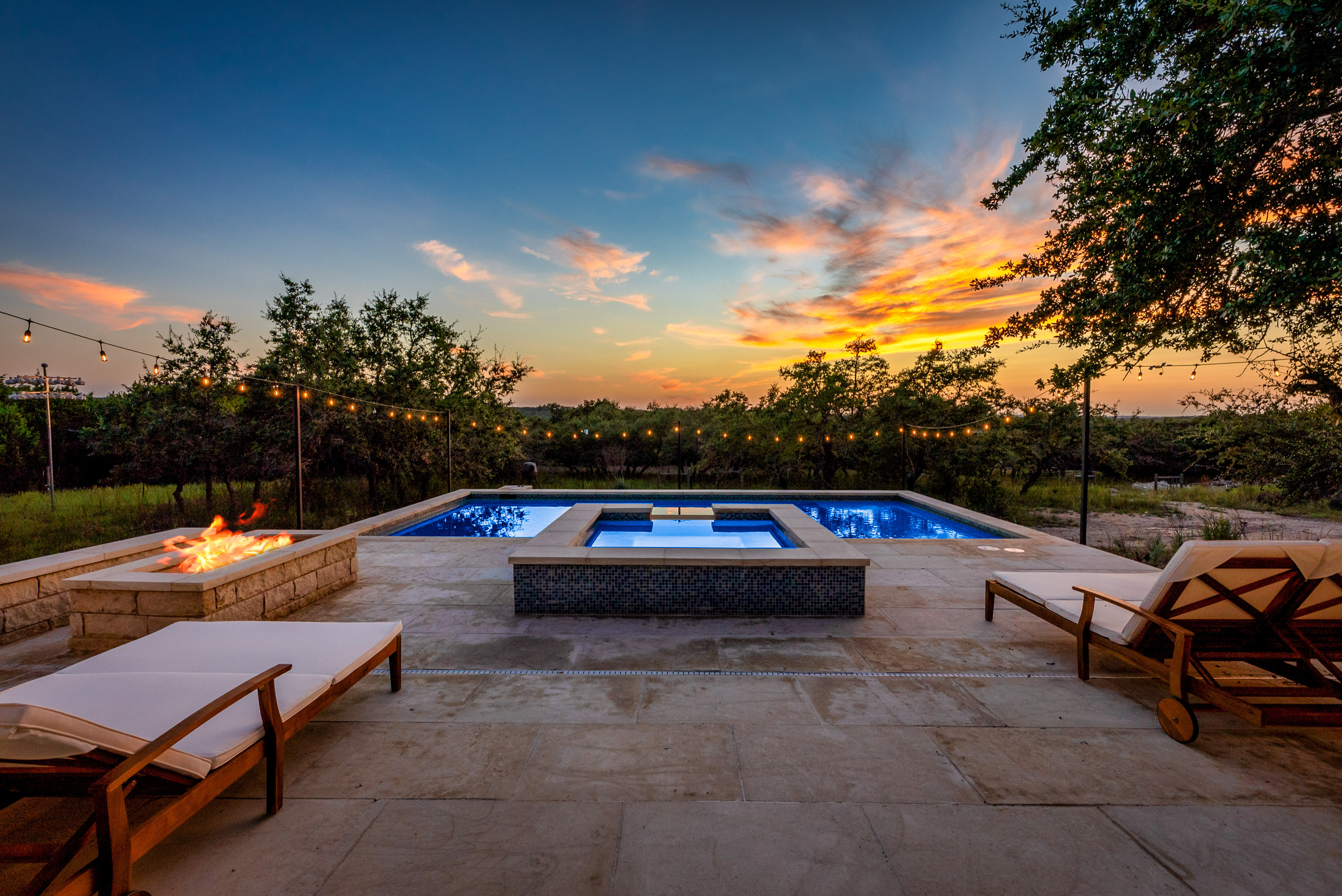 Twilight backyard pool and spa on a stone patio with a glowing fire pit, string lights, and wooden lounge chairs overlooking a treed landscape at sunset.
