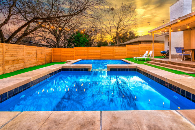 Suburban backyard swimming pool with attached spa at sunset, glowing blue water, wooden privacy fence, string lights and patio loungers