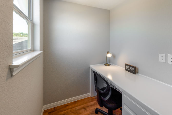 Compact sunlit home office nook with a white built-in desk, black mesh swivel chair, lit brass desk lamp and a small “Not now” decorative sign beside a window overlooking rooftops.