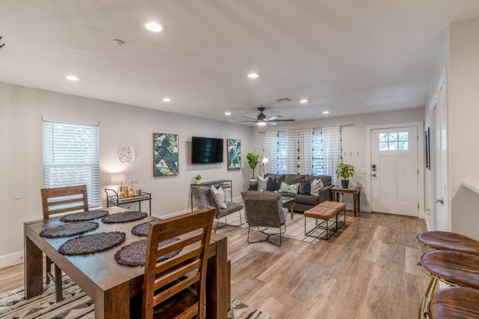 Cozy open-concept living and dining area with wood-look floors, dark wooden dining table and placemats, gray sofa and armchairs, wall-mounted TV, ceiling fan, potted plants, patterned curtains and a white entry door — modern neutral decor.