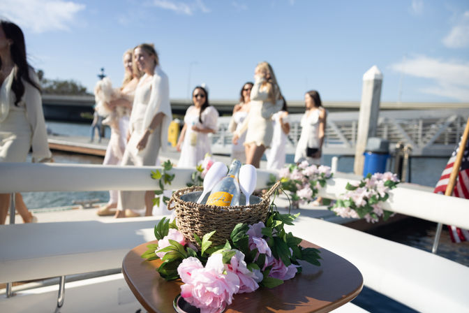 Woven basket holding a champagne bottle and glasses on a flower-decorated boat table, blurred women in white strolling at a sunny marina dock.