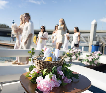 Woven basket holding a champagne bottle and glasses on a flower-decorated boat table, blurred women in white strolling at a sunny marina dock.