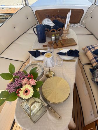 Cozy onboard dining nook on a boat with a table set for two — navy napkins, gold flatware, fresh floral bouquet, lantern and wooden candleholders on cream cushions