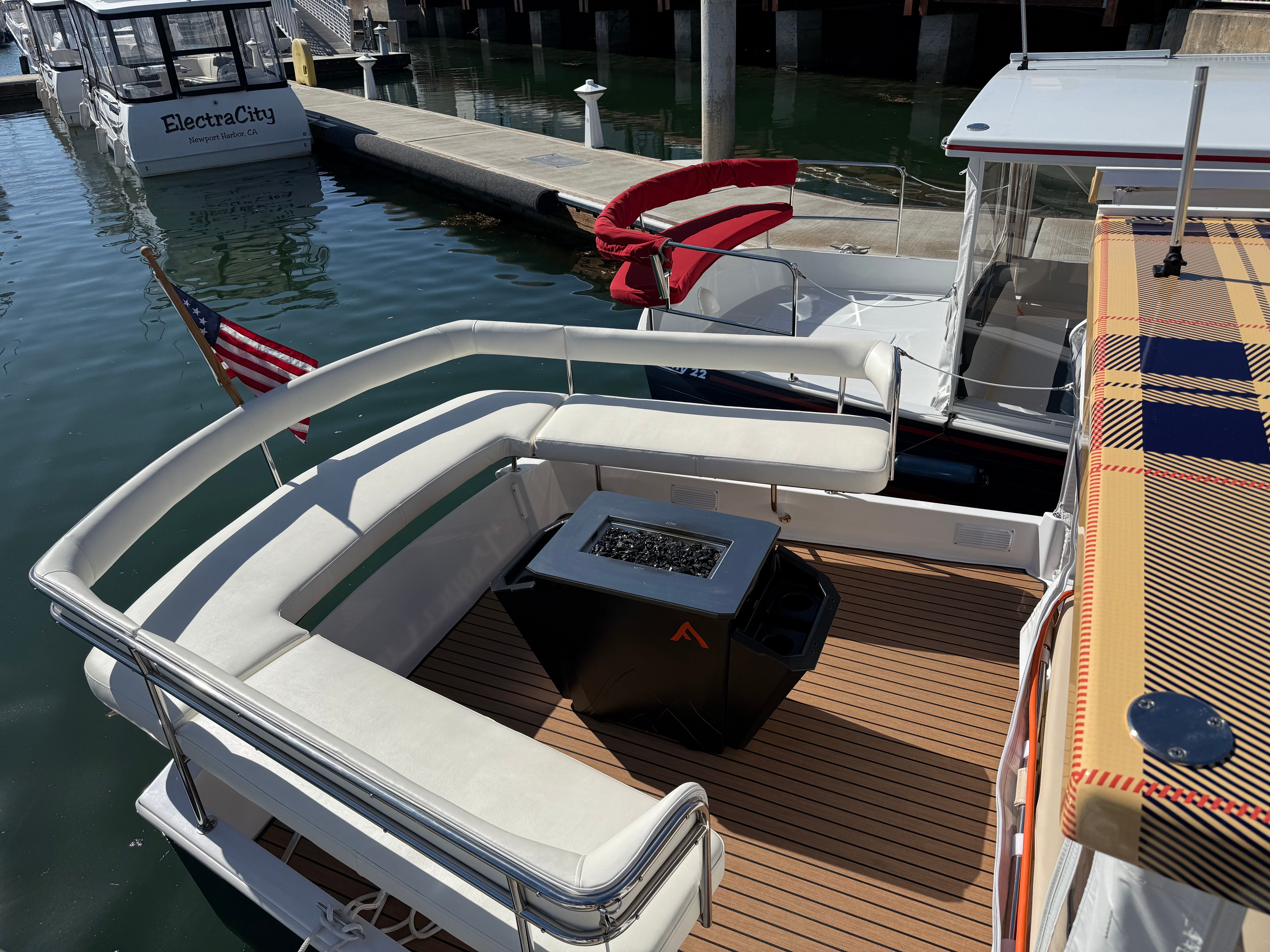 Sunlit yacht deck at a marina with white cushioned seating, teak-style flooring, a black firepit table, red curved boarding cushions, American flag at the stern and calm dockside water.