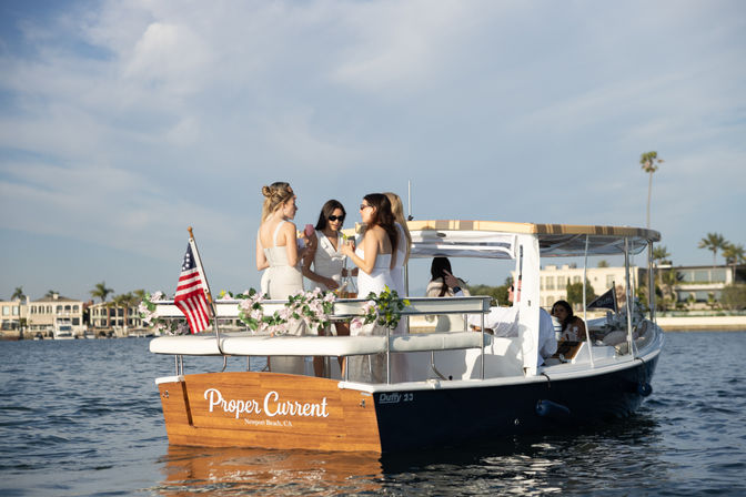 Four women in white dresses enjoying drinks on a floral-decorated boat near waterfront homes and palm trees, sunny blue-sky scene in Newport Beach, CA.