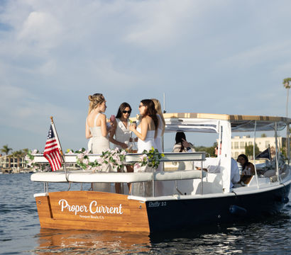 Four women in white dresses enjoying drinks on a floral-decorated boat near waterfront homes and palm trees, sunny blue-sky scene in Newport Beach, CA.
