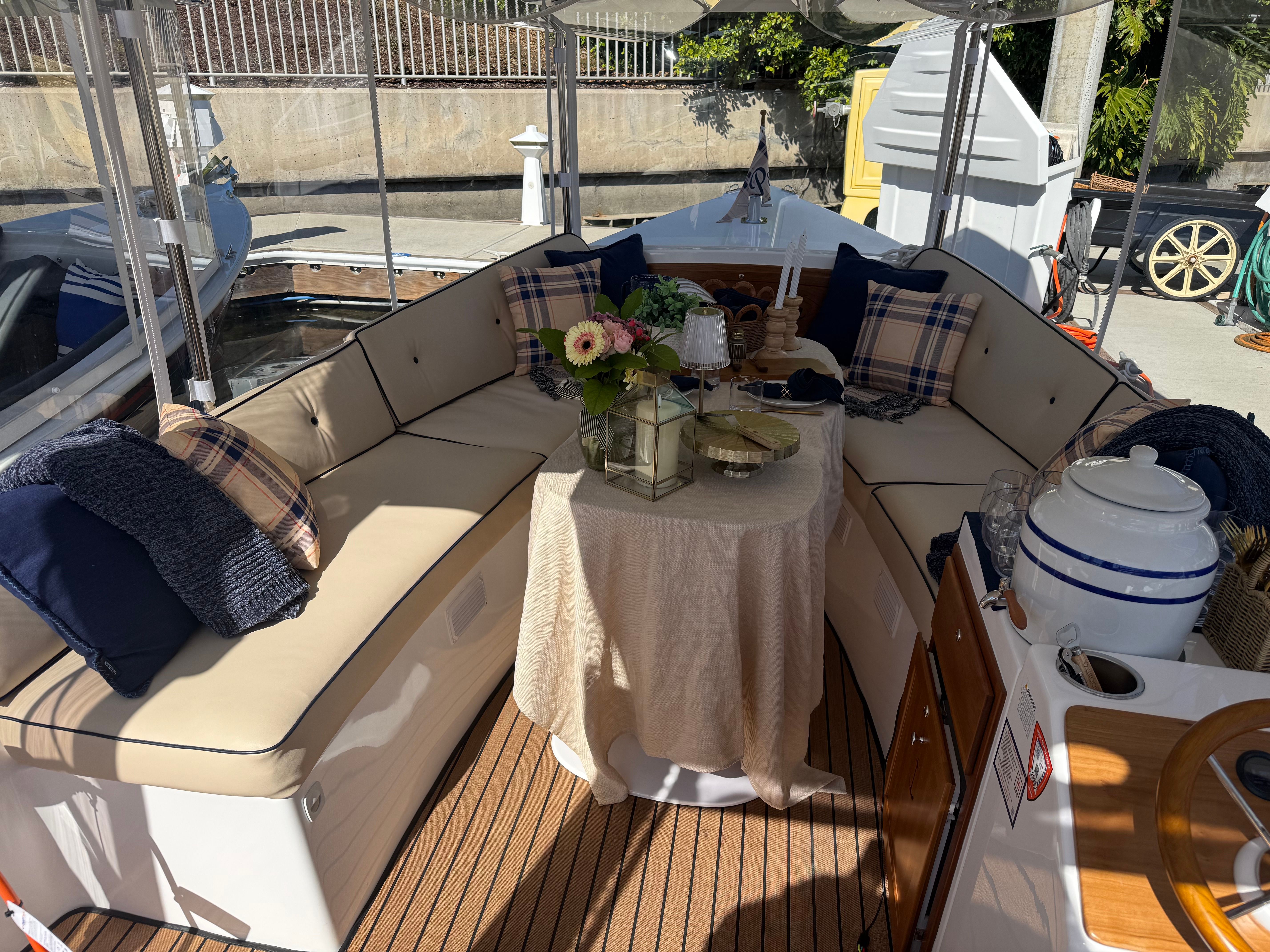 Cozy boat bow lounge with beige cushioned seating and navy plaid pillows, round table draped in linen with flowers, lantern and candles, teak deck and marina dock visible through clear enclosure