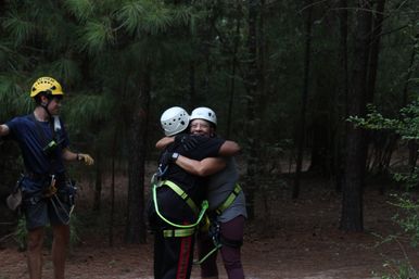 Two people in climbing helmets and safety harnesses share a joyful hug on a wooded ropes-course trail while a guide in a yellow helmet stands nearby among pine trees.
