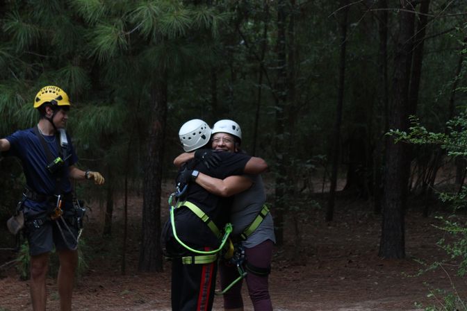 Two people in climbing helmets and safety harnesses share a joyful hug on a wooded ropes-course trail while a guide in a yellow helmet stands nearby among pine trees.
