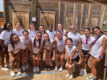 Smiling group of women in matching white event shirts posing at an outdoor axe-throwing range in front of wooden target boards and fenced lanes — team outing photo.