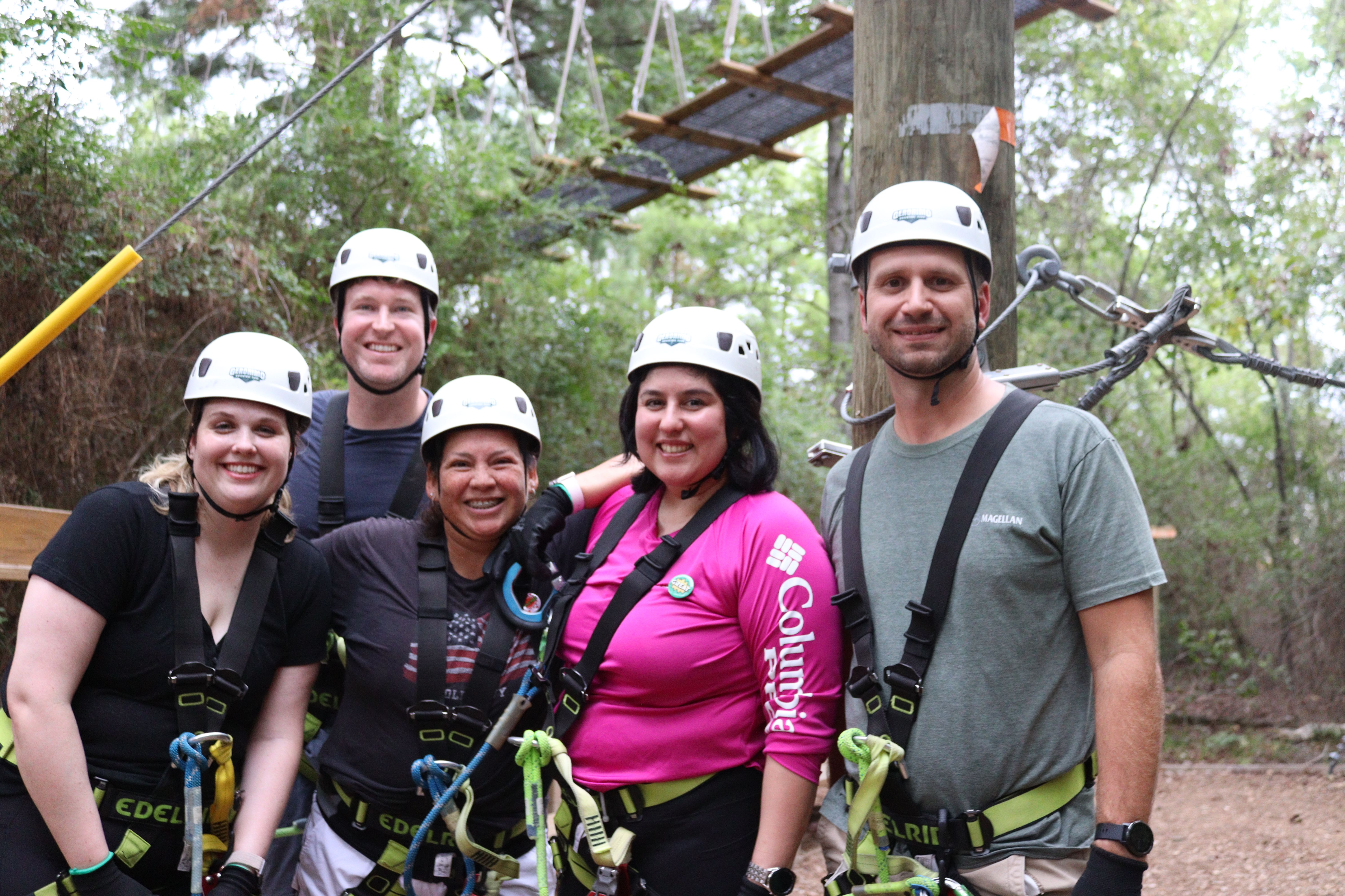 Five smiling adults in helmets and safety harnesses pose on a forest ropes course platform, ready for a zipline adventure