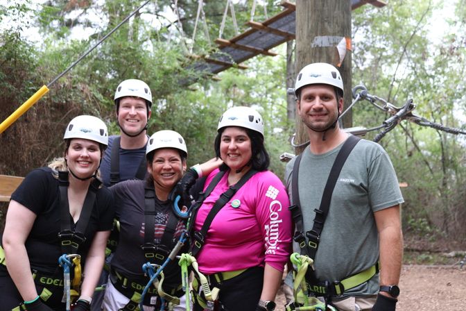Five smiling adults in helmets and safety harnesses pose on a forest ropes course platform, ready for a zipline adventure