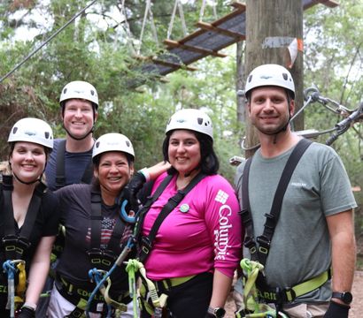 Five smiling adults in helmets and safety harnesses pose on a forest ropes course platform, ready for a zipline adventure