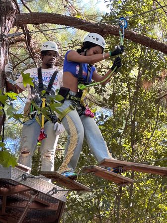 Two participants in helmets and harnesses navigating a wooden-plank ropes course high in a sunlit tree canopy, clipping safety lines on an outdoor adventure.